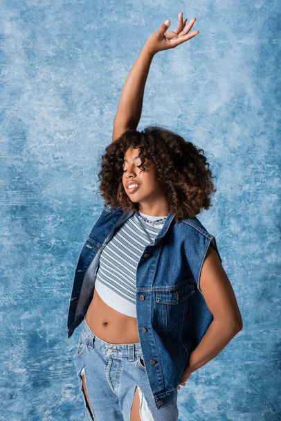 african american woman in denim vest and crop top standing with closed eyes and raised hand on blue background - Фото, изображение