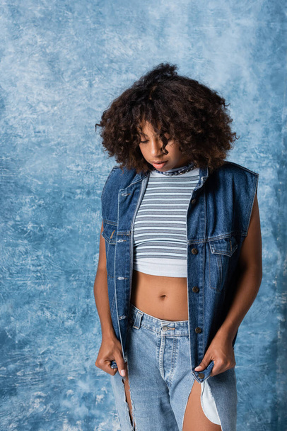 brunette african american woman in striped crop top and denim vest on blue background - Photo, Image