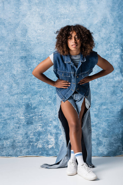 african american woman in ripped jeans and denim vest posing with hands on waist on blue background - Fotoğraf, Görsel