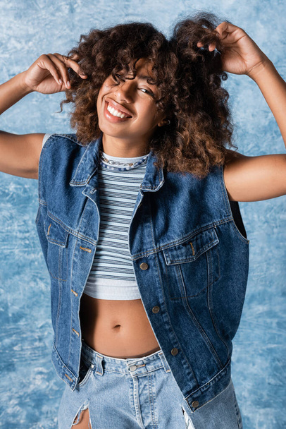 happy african american woman in denim vest touching wavy hair and looking at camera on blue background - Foto, Imagen