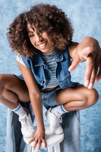 happy african american woman in denim clothes pointing at camera while sitting on haunches on blue background - Foto, Bild