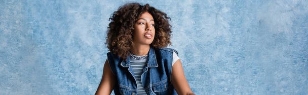 curly african american woman in denim vest looking away on blue background, banner - Photo, image