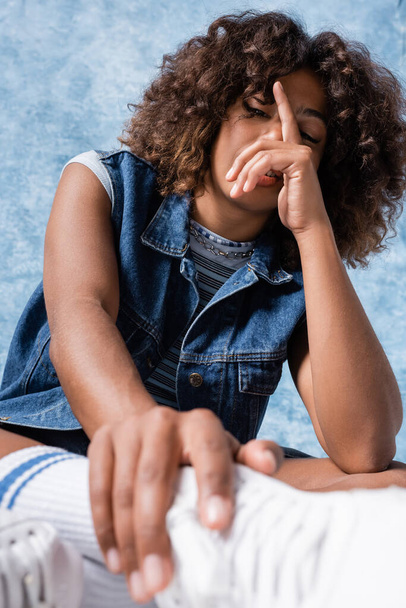 brunette african american woman in denim vest obscuring face with hand on blue background - Foto, imagen