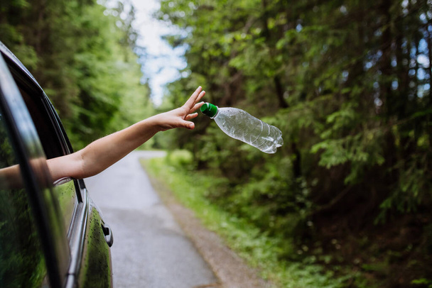 A womans hand throwing away plastic bottle from car window on the road in green nature, environmental protection, global warming concept - Foto, Imagen