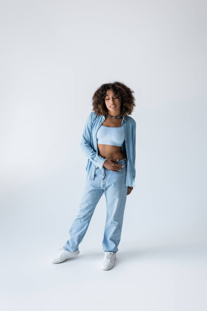 full length of african american woman in white sneakers and blue denim clothing on grey background - Foto, immagini