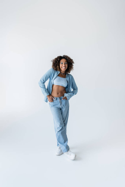 full length of pleased african american woman standing with hands in pockets of blue jeans on grey background - Foto, afbeelding