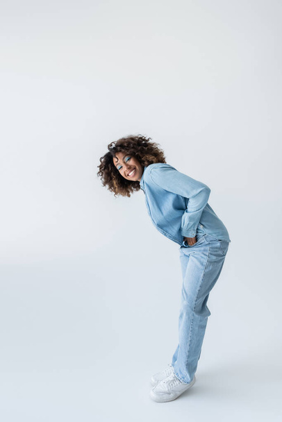 full length of happy african american woman in denim clothes standing with hand in pocket on grey background - Photo, Image