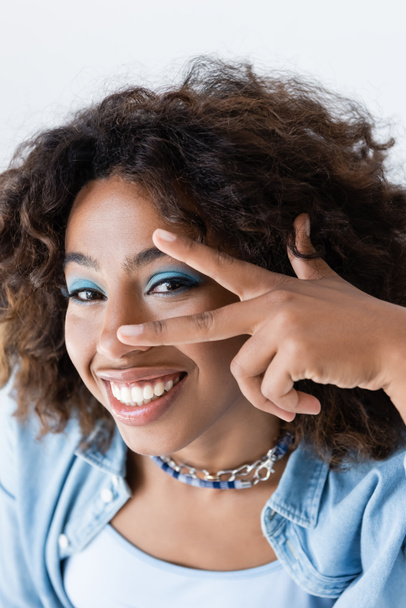 smiling african american woman showing victory sign near face with makeup isolated on white - Photo, image