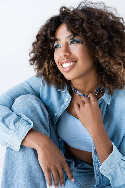 pleased african american woman in denim shirt touching necklaces and looking away isolated on grey - Фото, зображення