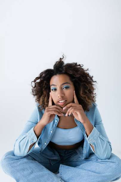 curly african american woman in denim clothes touching face and looking at camera isolated on grey - Valokuva, kuva