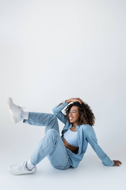 cheerful african american woman in denim clothes touching wavy hair while sitting on grey background - Fotografie, Obrázek