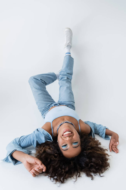 high angle view of happy african american woman in denim clothing lying on grey background - Fotó, kép