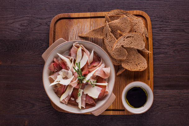 Prosciutto jerky in thin slices with parmesan and rosemary in a ceramic plate with handles. The plate is on a wooden board, next to it are rye bread chips and a saucepan with balsamic sauce. The board stands on a wooden background. - Foto, imagen