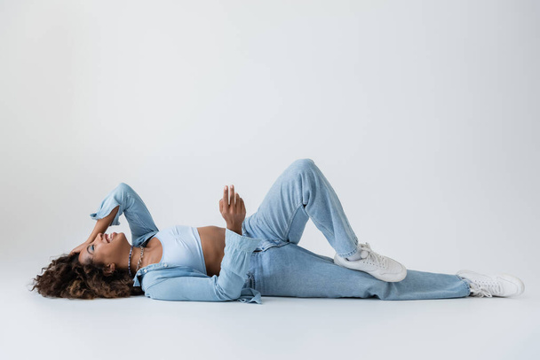 excited african american woman in blue denim clothes lying and laughing on grey background - Foto, Bild