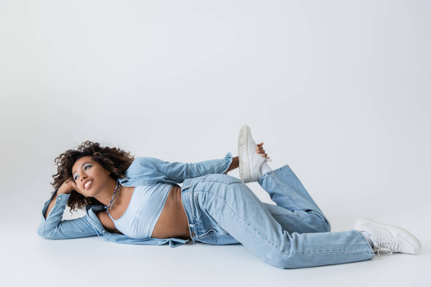 african american woman in trendy denim clothing looking away while lying on grey background - Fotografie, Obrázek