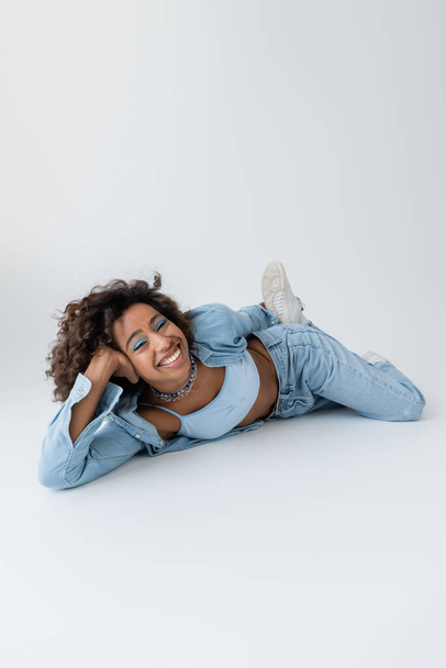 excited and stylish african american woman in denim clothing lying on grey background - Foto, imagen