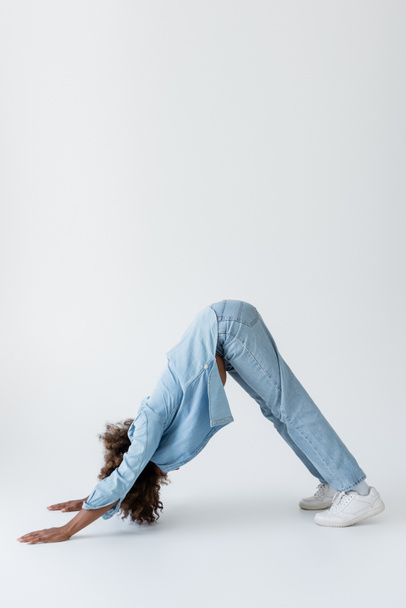 side view of african american woman in denim clothes standing in dolphin pose on grey background - Zdjęcie, obraz