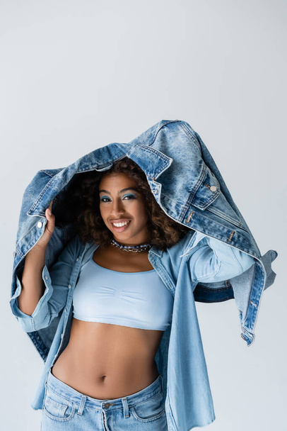 smiling african american woman in white crop top posing with denim jacket isolated on grey - Foto, afbeelding