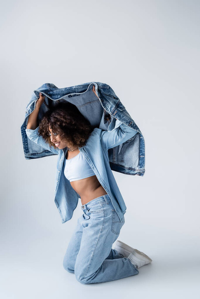happy african american woman posing with denim jacket while standing on knees on grey background - Foto, Imagen