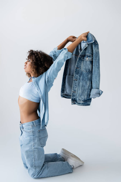 side view of african american woman holding denim jacket while standing on knees on grey background - Фото, изображение