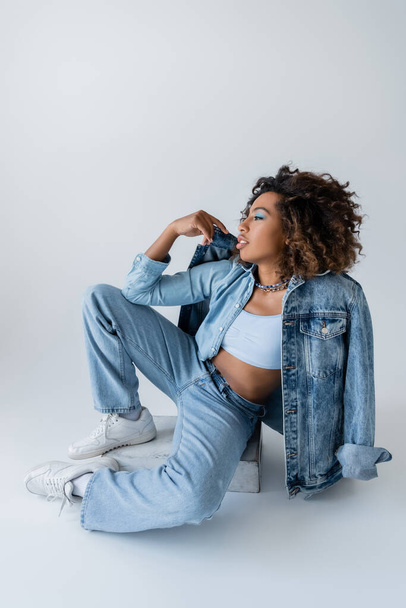 full length of stylish african american woman in white sneakers and blue denim clothes sitting on grey background - Foto, immagini