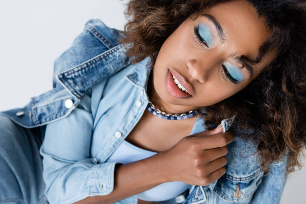sensual african american woman with blue eye shadows posing with denim garments isolated on grey - 写真・画像