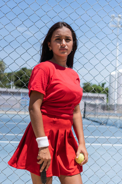 Jonge vrouw met racket en bal staan in de buurt van tennisbaan hek tijdens de pauze in de training. - Foto, afbeelding