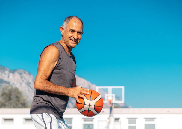 Half-length portrait of 60-years old smiling basketball player in good shape ready to shoot or pass a sport ball outdoor - Fotó, kép