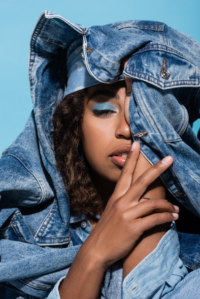 african american woman with makeup obscuring face with denim jacket isolated on blue - Photo, Image