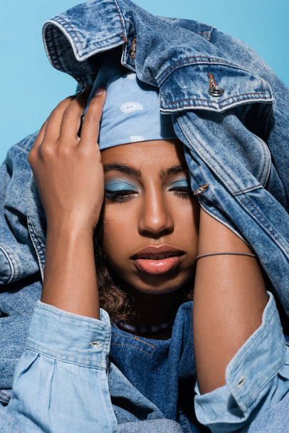 african american woman with makeup covering head with denim jacket isolated on blue - Fotografie, Obrázek