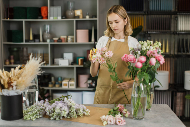 Woman florist at her own floral shop taking care of flowers. People, business, sale and floristry concept. Happy smiling florist woman making violet flower bunch at flower shop - Фото, изображение