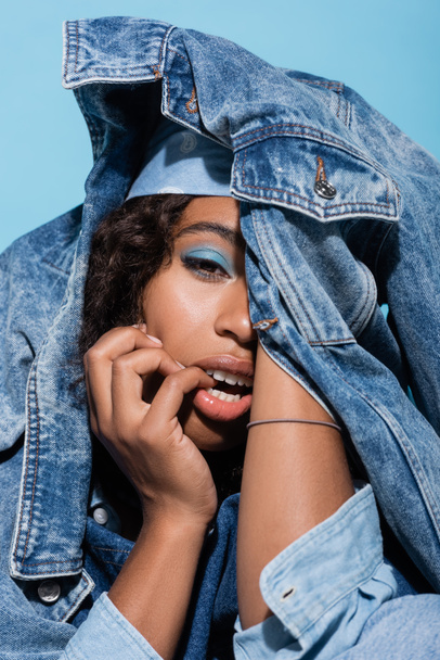 sensual african american woman obscuring face with denim jacket isolated on blue - Фото, зображення