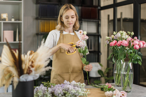 Cute girl florist collects a bouquet of roses in a flower shop. A beautiful florist creates a composition of flowers. Girl cuts flowers and removes stale leaves. - Foto, imagen