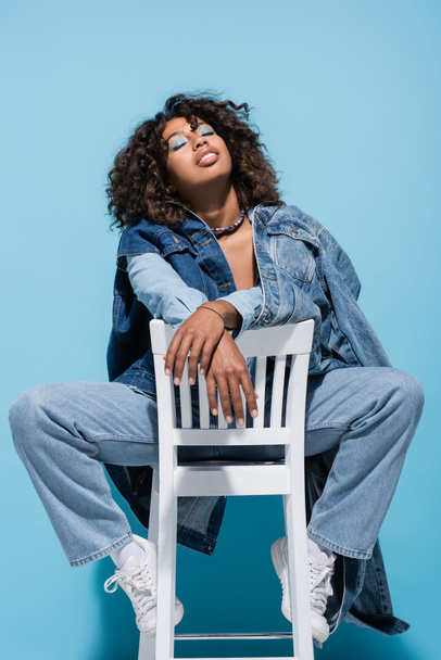 trendy african american woman in denim clothes posing on chair with closed eyes on blue background - Foto, imagen