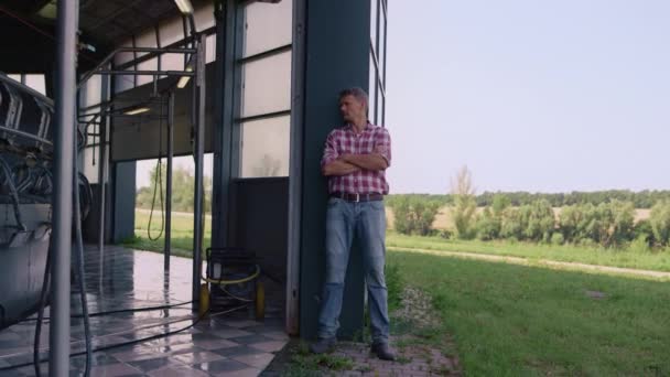 Farmer inspecting milk parlour at countryside. Tired worker rest lean facility wall. Thoughtful man agribusiness owner observing dairy production equipment on sunny day. Farming business concept. - Felvétel, videó