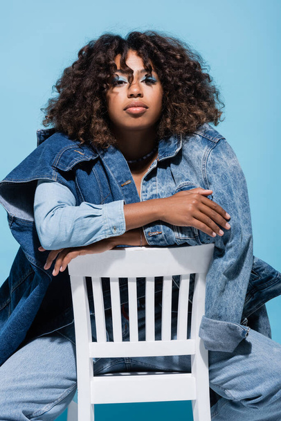 stylish african american woman with wavy hair sitting on chair with crossed arms isolated on blue - Foto, immagini