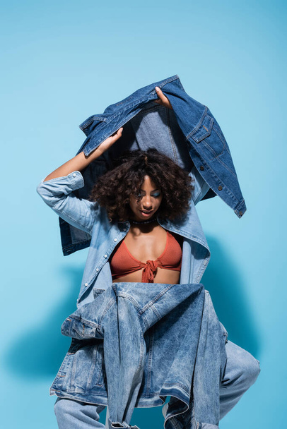 trendy african american woman holding denim jacket and vest while sitting on blue background - 写真・画像