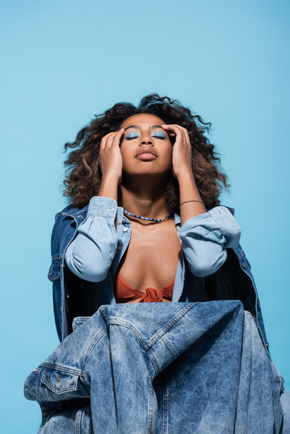 african american woman with makeup touching face while posing with denim clothes isolated on blue - 写真・画像