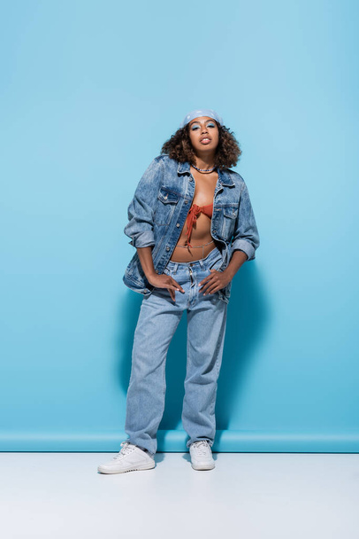 full length of african american woman in denim clothes posing with thumbs in pockets on blue background - Foto, immagini