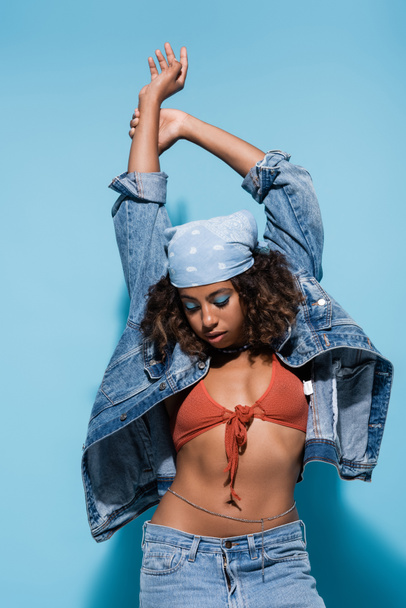 african american woman in swim bra and denim jacket standing with arms up on blue background - Φωτογραφία, εικόνα