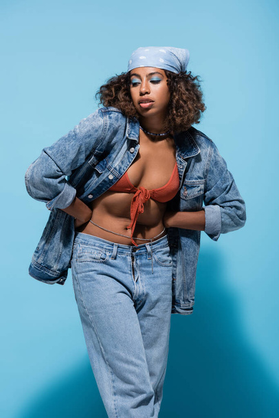sensual african american woman in denim clothes and swim bra standing with hands on waist on blue background - Fotografie, Obrázek