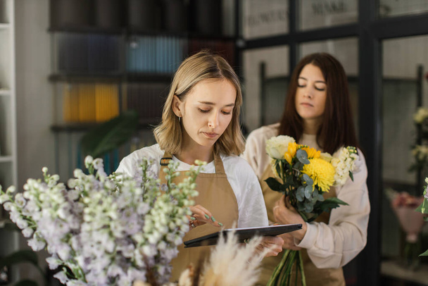 Creative female designer sit at desk in modern shop interior and make order for next period, woman assistant compose arrange plants flowers in bouquets and make floral compositions for sale. - 写真・画像