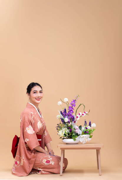 Young 20s Asian Japanese woman wear Pink traditional Kimono, does Ikebana flower arrangement. Japan Kimono female express felling happy smile over pastel yellow background isolated copy space - Фото, изображение