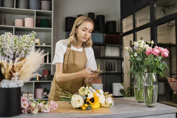 Caucasian female florist making bouquet at work in floral shop and taking pictures. Young pretty woman taking photo of finished flowers bouquet for social media. Floristry concept. - 写真・画像