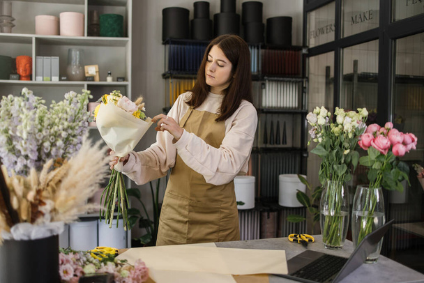 Portrait of young brunette female florist holding bouquet of flowers and looking at them. Professional and hardworking florist. - Zdjęcie, obraz