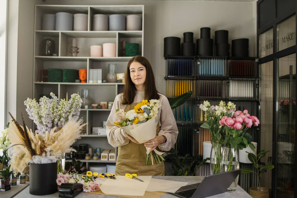 Smiling florist in uniform work in a flower shop behind counter with different varieties of flowers and huge bouquet in hands. Small business concept. Modern loft interior. - Fotoğraf, Görsel
