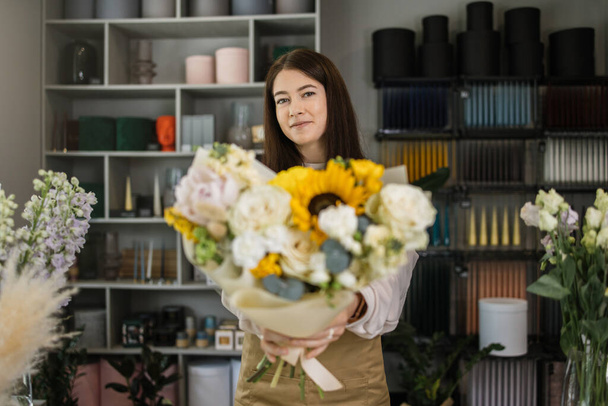 Smiling florist in uniform work in a flower shop behind counter with different varieties of flowers and huge bouquet in hands. Small business concept. Modern loft interior. - Valokuva, kuva