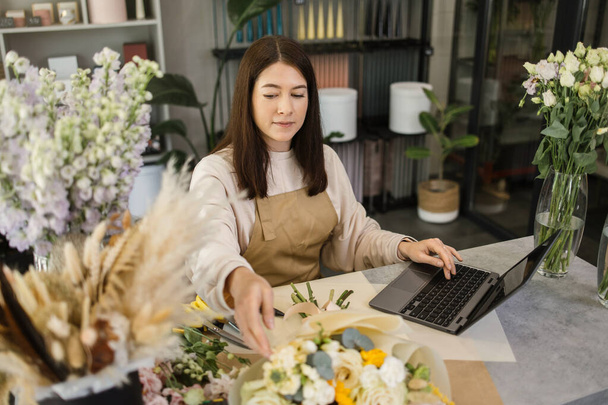 Portrait of attractive woman florist working on computer at workplace. Concentrated female sitting at counter in floral store and typing on laptop. - Photo, Image