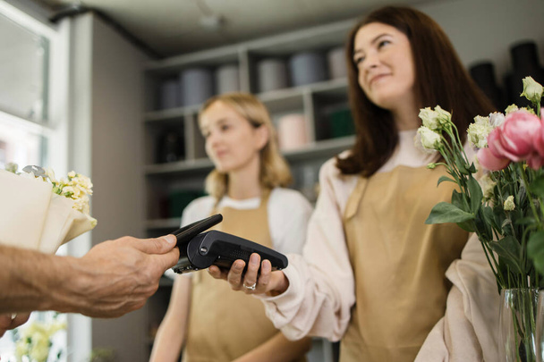 Two smiling woman florists holding card reader machine at counter while customer paying with electronic credit card in his smartphone. - Photo, Image