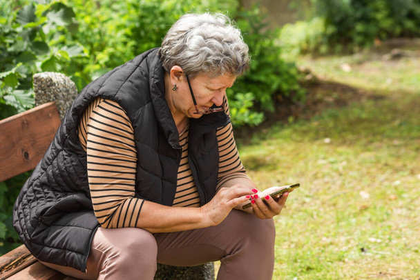 Boční pohled na kavkazskou starší ženu s brýlemi sedícími na lavičce v parku a používající smartphone. Šedovlasá dáma v neformálním oblečení si povídá na sociálních sítích a užívá si mobilní služby. Starší koncepce života. - Fotografie, Obrázek